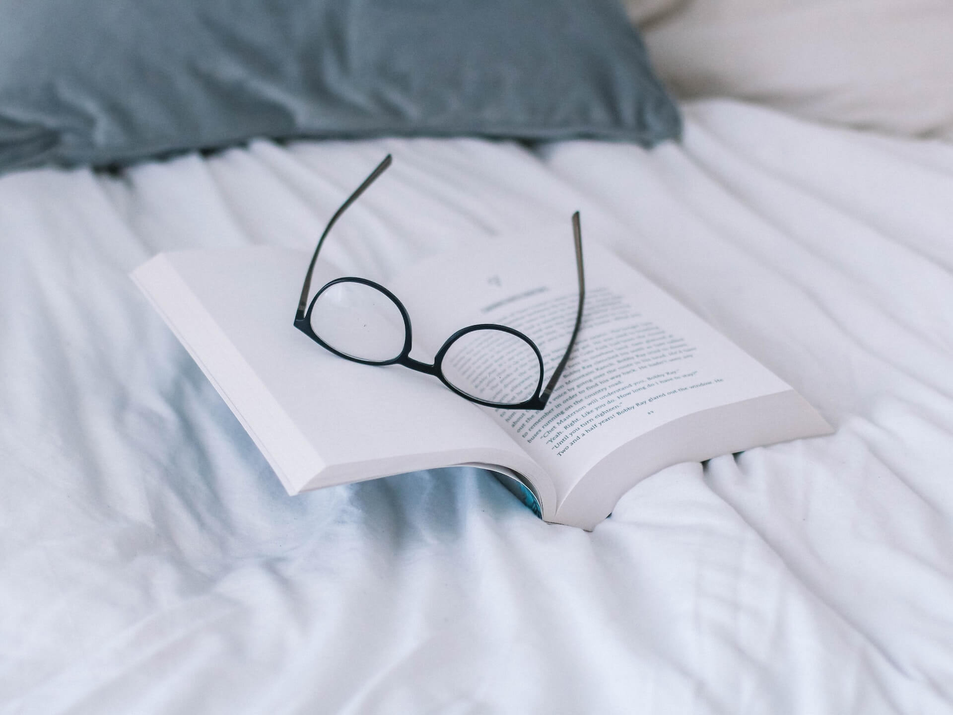 An open paperback book on a bed with a pair of glasses resting between the pages