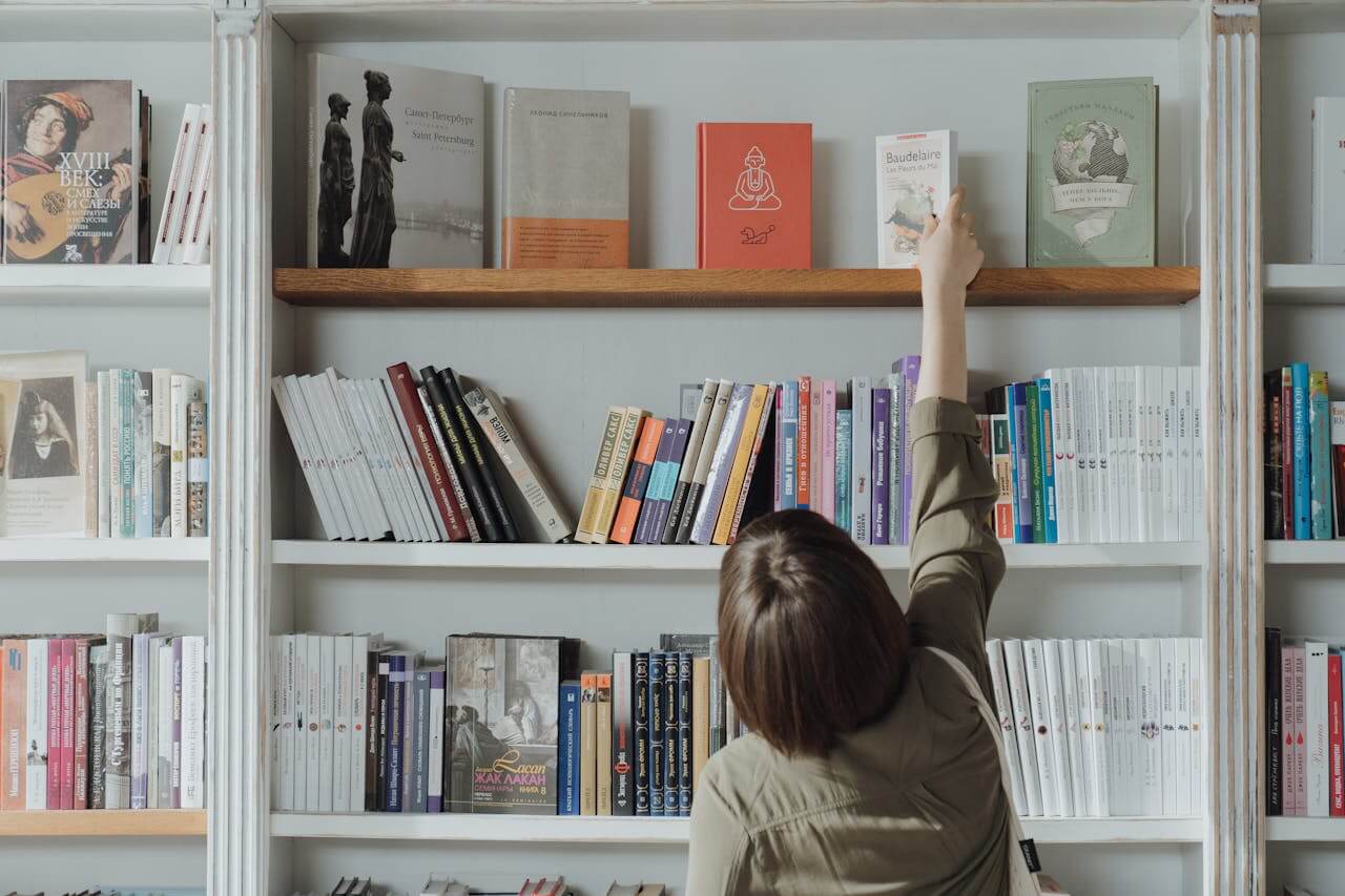 A woman stands in front of a bookshelf and reaches up to a book on the top shelf.